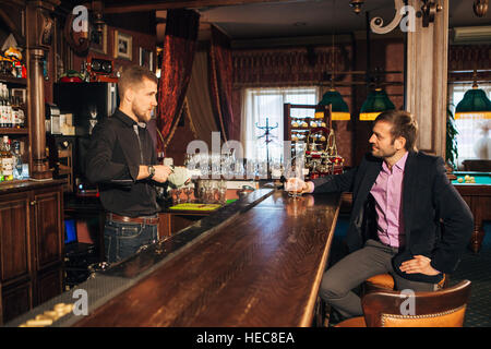 Sorridente attraente giovane barista tergi bicchieri e a parlare con l'uomo in bar Foto Stock