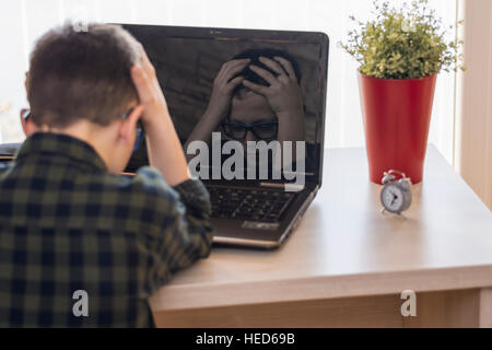 Sconvolto il ragazzo con gli occhiali utilizzando computer portatile mentre è seduto sulla scrivania a casa. Viso espressivo in riflessione. Foto Stock