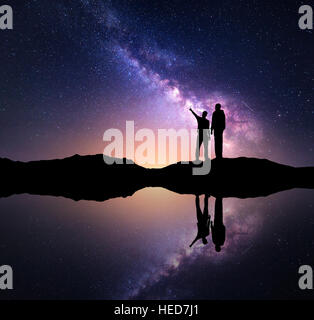 La Via Lattea con la silhouette di una famiglia. Padre e figlio che puntare il dito nella notte cielo stellato in montagna vicino al lago Foto Stock