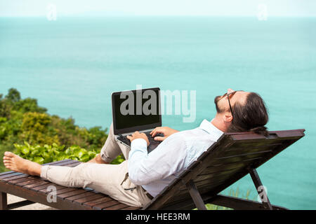 Imprenditore indossa una tuta sulla sedia a sdraio digitando portatile in spiaggia Foto Stock
