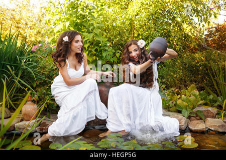 Due sorelle in vestiti bianchi presso il laghetto di ninfee. La ragazza si versa acqua da piccoli vasi di creta torna nello stagno. Foto Stock