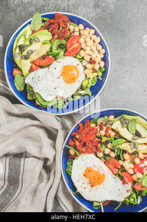 Sana colazione ciotole con uovo fritto, germogli di ceci, semi e verdure fresche e verdi su cemento grigio Sfondo, vista dall'alto. Pulire mangiare, di Foto Stock