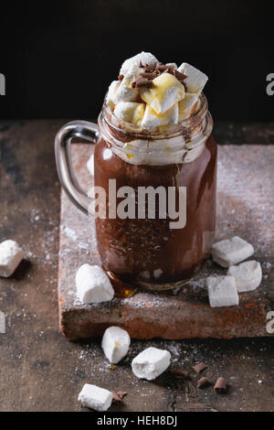 La cioccolata calda con marshmallow casalinga, scaglie di cioccolato e sciroppo di mason jar, in piedi sul bordo di terracotta su un vecchio legno scuro dello sfondo. Spazio f Foto Stock