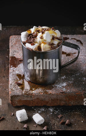 La cioccolata calda con marshmallow casalinga, scaglie di cioccolato e sciroppo nella tazza di metallo, in piedi sul bordo di terracotta su un vecchio legno scuro dello sfondo. Copia sp Foto Stock