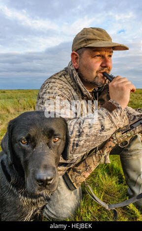 Un Regno Unito wildfowler, o cacciatore di anatre, sat sul foreshore o marsh con il suo cane soffiando una chiamata anatra in lo sbiadimento della luce della sera Foto Stock