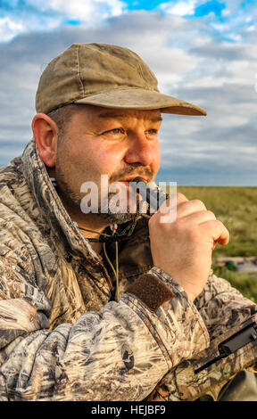 Un Regno Unito wildfowler, o cacciatore di anatre, sat sul foreshore o marsh soffiando una chiamata anatra in lo sbiadimento della luce della sera Foto Stock