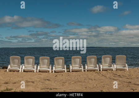 Una fila di otto sedie in plastica bianca allineati su di una spiaggia di sabbia di fronte ad un lago Foto Stock