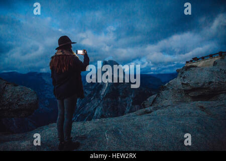 Giovane donna prendendo fotografia su roccia affacciato sul Parco Nazionale di Yosemite al crepuscolo, CALIFORNIA, STATI UNITI D'AMERICA Foto Stock