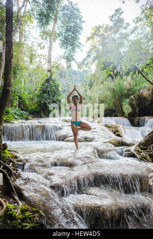 Donna in piedi su una gamba sola, nella struttura ad albero pongono, in cascate di Kuang Si, Luang Prabang, Laos Foto Stock