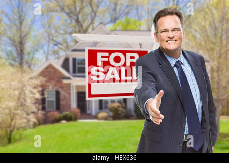 Sorridente agente maschile raggiungendo per un tremolio della fotocamera nella parte anteriore della bella casa e per la vendita immobiliare di segno. Foto Stock