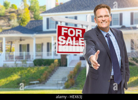 Sorridente agente maschile raggiungendo per un tremolio della fotocamera nella parte anteriore della bella casa e per la vendita immobiliare di segno. Foto Stock