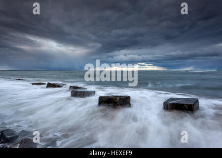 Cubi di calcestruzzo mezzo sepolto nella sabbia in spiaggia Cresswell, sulla costa di Northumberland al tramonto ad alta marea, tempesta in arrivo Foto Stock