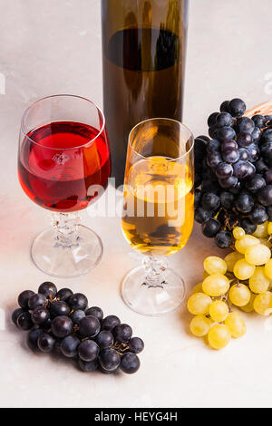 Due bicchieri - uno con il vino bianco e uno con un bicchiere di vino rosso e marrone con bottiglia di vino bianco sul marmo chiaro dello sfondo. Grappoli di uva Foto Stock