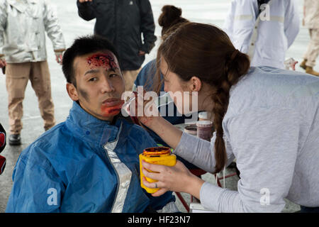 Seiya Chinen, sinistra, ha sangue artificiale e il trucco applicato, una tecnica nota come moulage, 24 Aprile presso il Naha porto militare. Durante l'esercizio, gli enti locali e la polizia militare, vigili del fuoco e del personale del servizio di emergenza eseguito una serie di allenamenti pianificati eventi in risposta a una simulazione di crash di un aereo militare in una zona urbana. Il trucco e gore è stata utilizzata per la fabbricazione di aeromobili Off-Base contrattempo Esercizio 2014 nel modo più realistico possibile. Chinen è un vigile del fuoco con la città di Naha dei Vigili del Fuoco. (U.S. Marine Corps foto di Cpl. Natalie M. Rostran/RILASCIATO) Giappone, noi primi responder treno Foto Stock