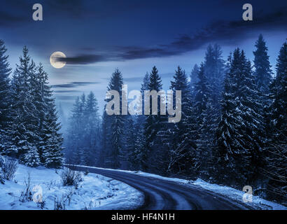 In inverno il paesaggio di montagna. tortuosa strada che conduce nel bosco di abete rosso con neve di notte nella luce della luna piena Foto Stock