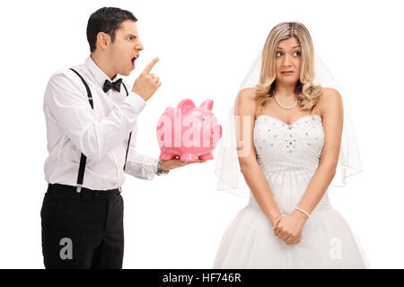 Arrabbiato groom tenendo un piggybank e scolding un imbarazzato sposa isolati su sfondo bianco Foto Stock