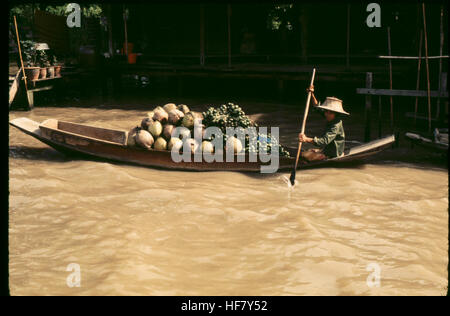 Questa immagine mostra una barca tradizionale a Bangkok, Thailandia, che galleggia lungo un canale, carica di cocco e frutta, mentre una donna che indossa un tipico cappello fila la barca. Foto Stock