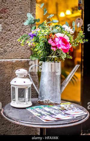 Un bellissimo colorato annaffiatoio pieno di fiori si siede sul lato della strada cafe tabella a Barcellona, Spagna. Foto Stock