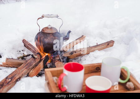 Inverno romantico picnic. Due coppe e una ciotola su un vassoio di legno nella neve. Bollitore di rame su un fuoco aperto su sfondo sfocato. Pentola su firewo Foto Stock