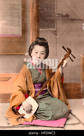 Circa 1900, seppia cartolina giapponese. Donna, geisha inginocchiati sul pavimento giocando shamisen. Contatto visivo. Colorate a mano. Foto Stock