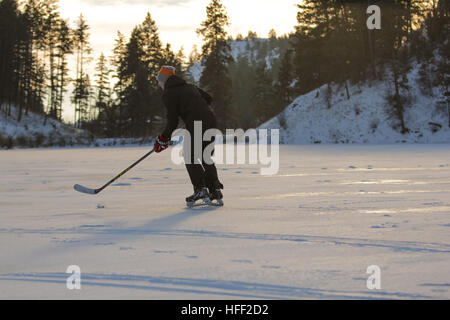 Ragazzo canadese che pattina su un lago ghiacciato giocando hockey su ghiaccio in un pomeriggio invernale soleggiato ma freddo Foto Stock