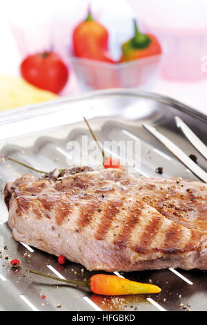 Carne di maiale alla griglia cotoletta, grill marchi, su una griglia in alluminio con forcella di carne, ciotola di peperoni in background Foto Stock