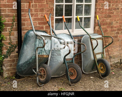 Fila di tre giardino metallo carriole appoggiata contro il vecchio rosso muro di mattoni, UK. Foto Stock