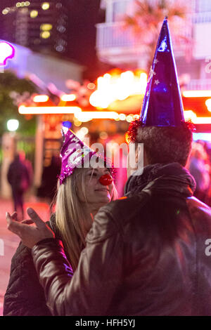 Benidorm, Spagna. Il 31 dicembre, 2016. I turisti e i locali, goditi la musica e le danze sul lungomare e la spiaggia. Coppia con naso rosso e ha fatto parte di cappelli di parlare di fronte a luci luminose Foto Stock