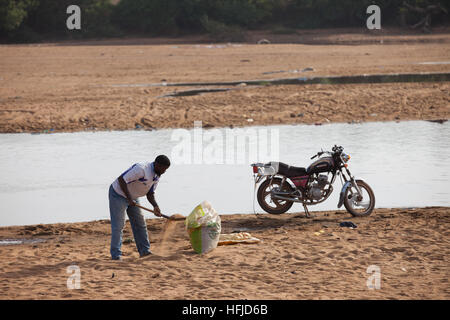 Kiniero, Guinea, 20 Aprile 2015: Un uomo raccoglie la sabbia dalla banca rivert al trasporto sulla sua moto. Foto Stock