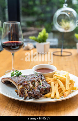 Grigliata di carne di maiale alla brace bimbo indietro nervature grigliate di mais dolce e patatine fritte sul tavolo da pranzo con un bicchiere di vino rosso Foto Stock