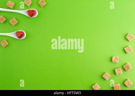 Due cucchiai di bianco con cuori rossi su di essi e lo zucchero di canna cubi su sfondo verde. Vista dall'alto. Copia spazio per il testo. Foto Stock