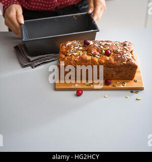 Una donna elegante mani tenere una teglia da forno su un banco di cucina. Accanto ad esso, un mirtillo palustre e seme fatta in casa pagnotta cotta è raffreddamento, riempimento la casa wi Foto Stock