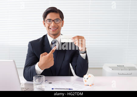 Giovani professionisti uomo mostra business card presso un ufficio con computer portatile e salvadanaio su tavola Foto Stock