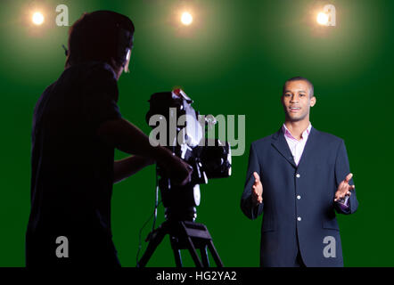 Il presentatore a parlare con la fotocamera in una schermata verde studio con stagliano cameraman in primo piano e le luci in studio in background. Foto Stock