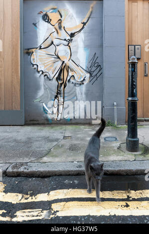 Un gatto grigio che attraversa la strada di Princelet Street nella zona est di Londra con un murale in background di una ballerina Foto Stock