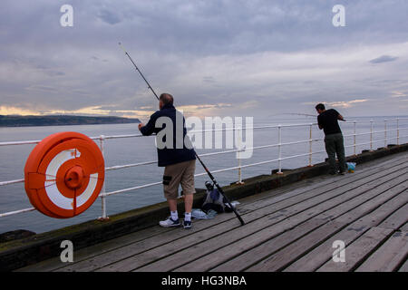 Stagliano contro un oscuro, nuvoloso, cielo di sera, due uomini sono la pesca in mare dal Molo Ovest - Whitby, North Yorkshire, Inghilterra. Foto Stock