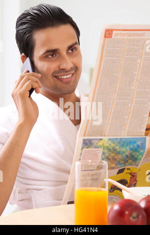Uomo che parla al telefono e leggere il giornale al tavolo per la colazione Foto Stock