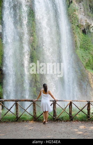 Giovani attraenti brunette donna che indossa un abito bianco, rilassata e serena in un bellissimo luogo naturale, di fronte ad una grande cascata, tra un assortimento di v Foto Stock