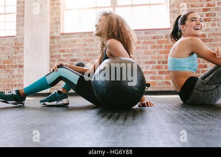 Felice giovane femmina amici seduti sul pavimento Palestra con palla medica e sorridente. Due giovani donne che prendono una pausa da allenamento. Foto Stock