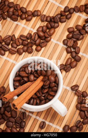 Tazza di caffè riempiti con i chicchi di caffè e due bastoncini di cannella su una stuoia di bambù con ornamenti di chicchi di caffè. Foto Stock