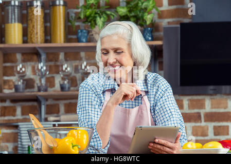 Senior sorridente donna in grembiule utilizzando digitale compressa in cucina Foto Stock