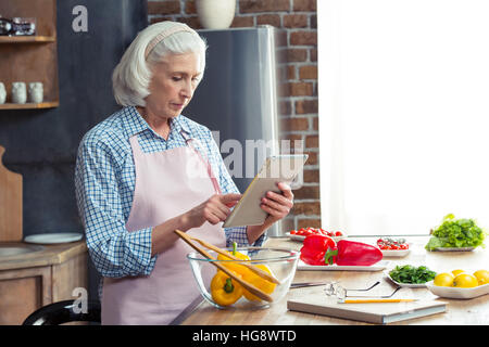 Donna senior con tavoletta digitale in cucina durante la preparazione di insalata di verdure Foto Stock