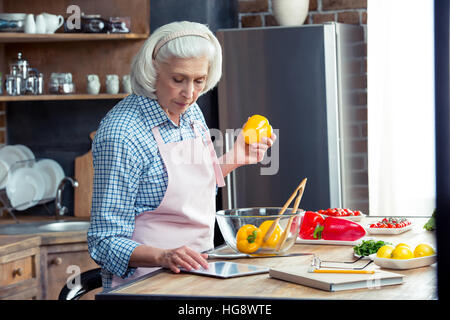 Donna senior con tavoletta digitale in cucina durante la preparazione di insalata di verdure Foto Stock