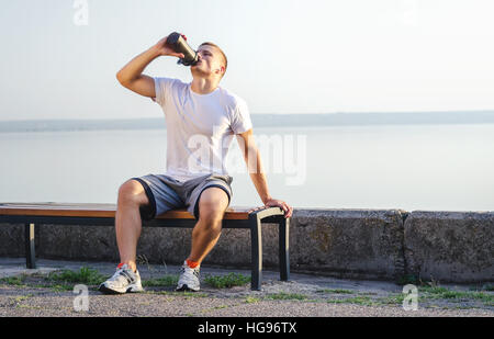 Giovane uomo sulla panchina dopo il jogging all'aperto. Atleta di bere da un agitatore al tramonto. Foto Stock