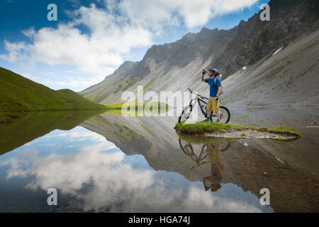 Un mountainbiker è acqua potabile mentre si sta in piedi in un bellissimo lago di montagna. La regione di Lenzerheide nei Grigioni in Svizzera è una singola via paradiso. Foto Stock