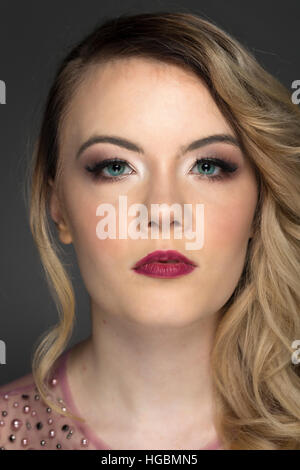 Colpo di Testa del modello con la bionda bouncy ricci, trucco naturale e profonda berry labbra rosa da indossare jeweled alto fondale grigio Foto Stock