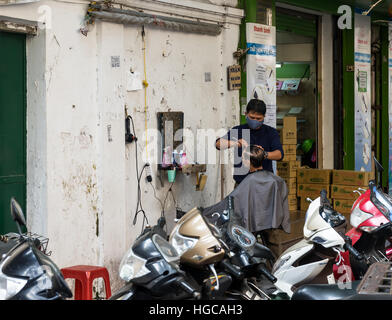 L'uomo il taglio di capelli al suo barbiere business per le strade di Hanoi, Vietnam. Il barbiere si trova sulla strada aperta. Foto Stock