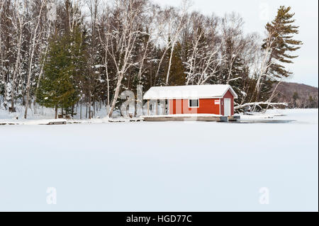 Paesaggio innevato canadese, rifugio rosso in un lago ghiacciato in inverno vicino a Huntsville, Ontario rurale, Ontario, Canada. Foto Stock