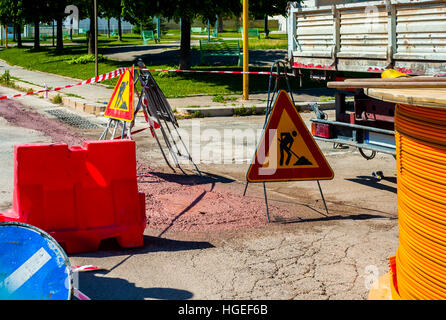 Cartelli di avvertimento per i lavori in corso sulla strada in costruzione. cavi a fibra ottica sepolto in un micro trench con calcestruzzo colorato di rosso da un lavoratore Foto Stock