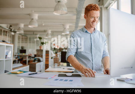 Colpo di felice giovane uomo in piedi alla sua scrivania e lavorare sul computer. Imprenditore lavora in un ufficio moderno. Foto Stock